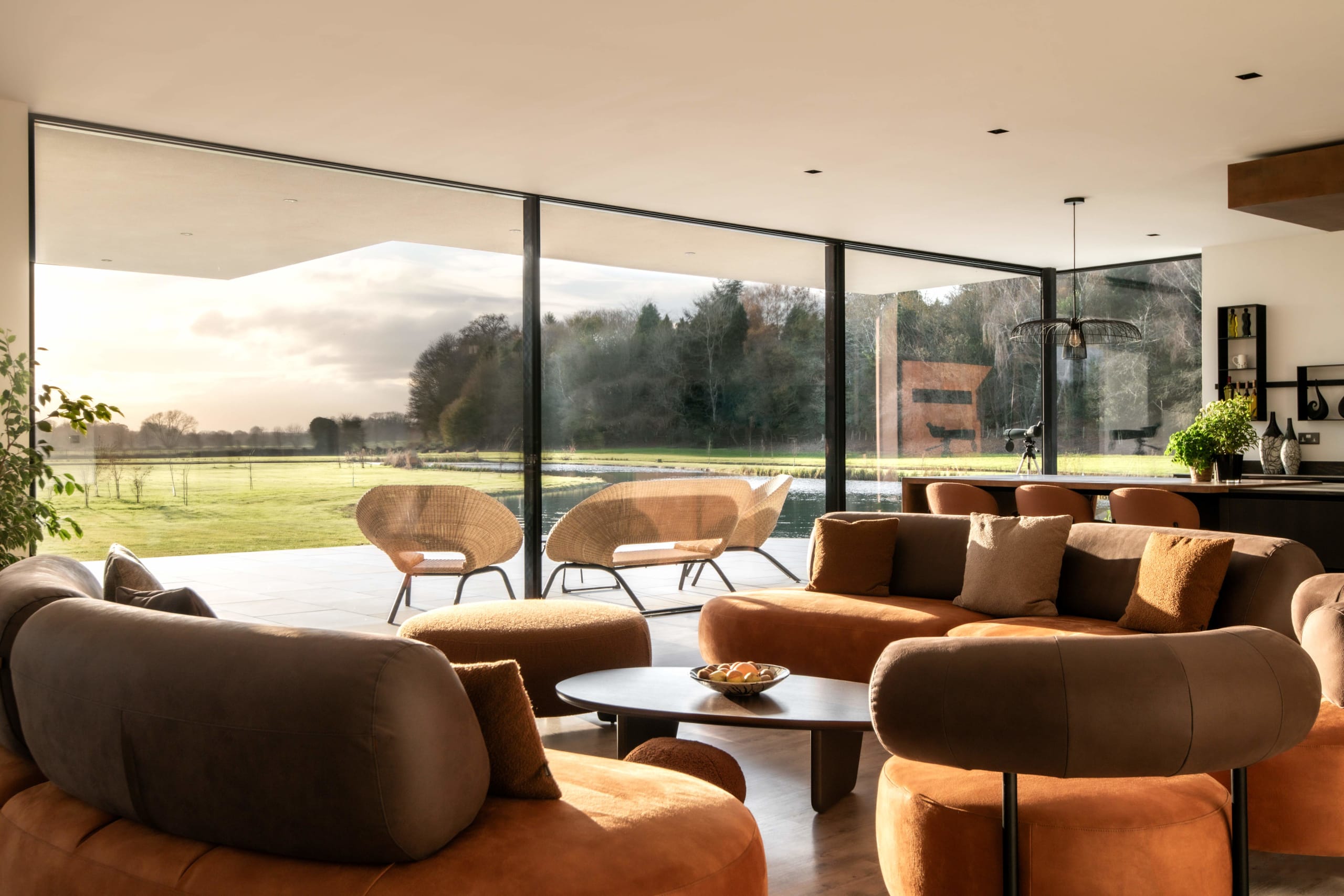 Modern living room with curved terracotta and taupe sofas, floor-to-ceiling windows overlooking countryside and pond